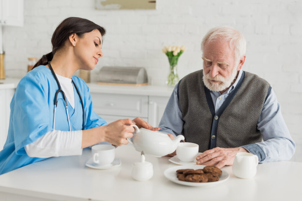 Nurse pouring tea for senior man, sitting on kitchen at home